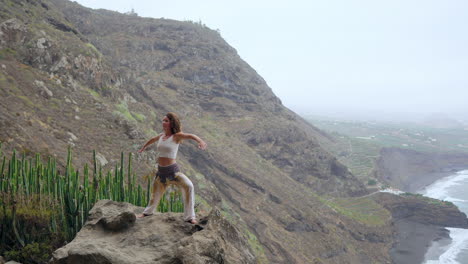 En-La-Costa-Cerca-De-Montañas-Rocosas,-Una-Mujer-Practica-La-Postura-Guerrera-Del-Yoga,-Meditando-En-Medio-De-La-Extensión-Del-Océano:-Un-Retrato-De-Motivación-E-Inspiración-En-El-Fitness.